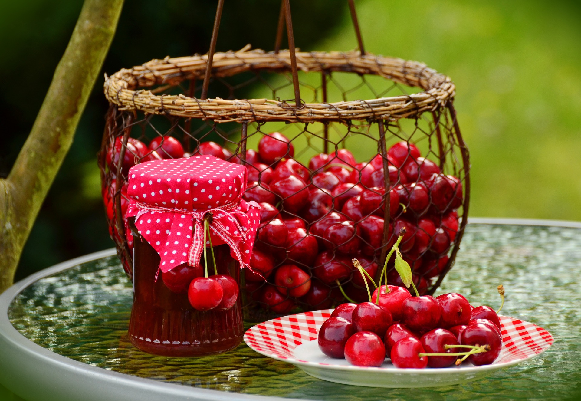 Basket of fresh cherries on small glass table witj jar of cherry jam and some cherries on a red-and-white plate.