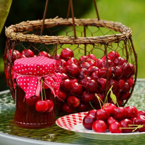 Jar of cherry jam standing next to a basket and plate of fresh cherries