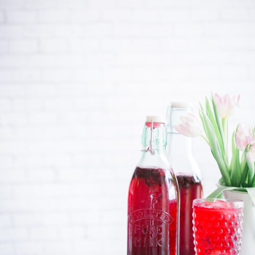 two bottles of raspberry cordial, with a glass of cordial in the foreground. On marble top.