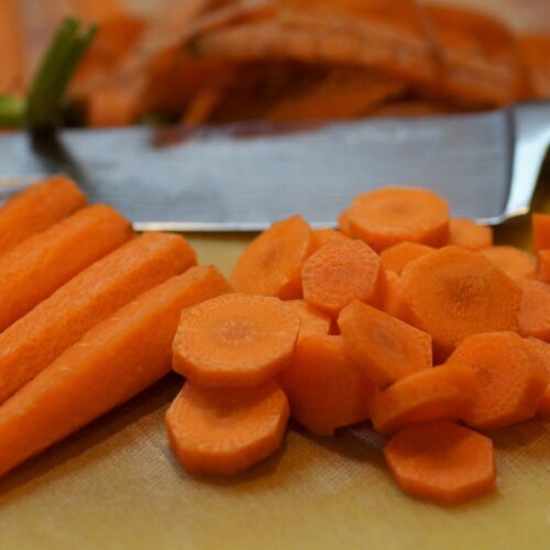 Chopping board with knife in background. Foreground has peeled carrots at the top and a pile of chopped carrot rounds in the middle