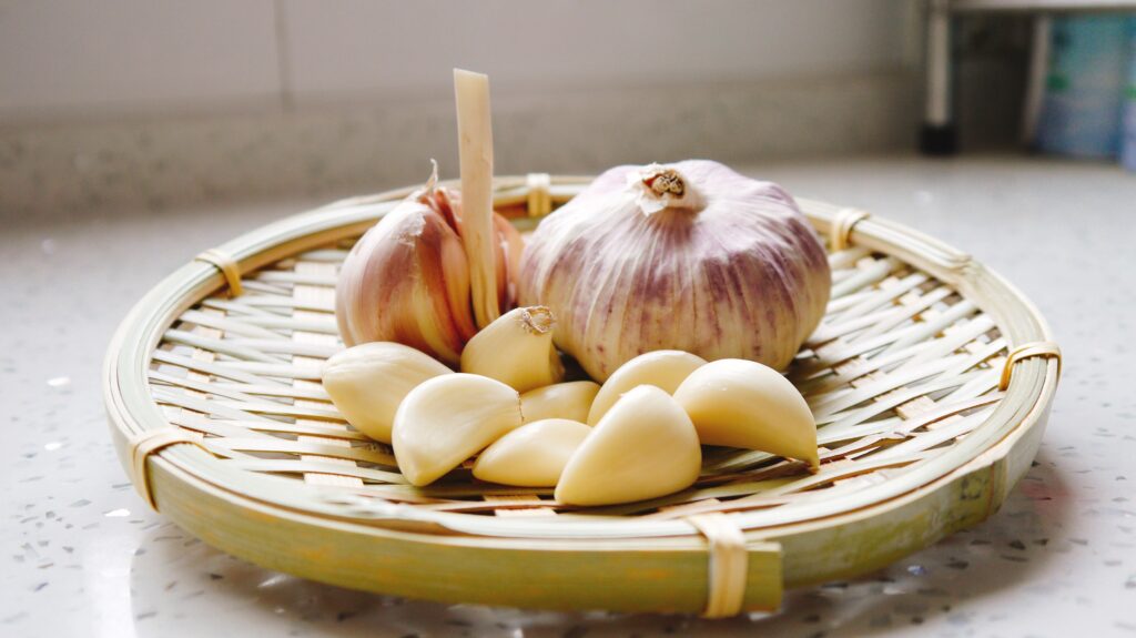 One whole and one half bulb of pinkish garlic, alongside a pile of peeled garlic cloves on a basket top