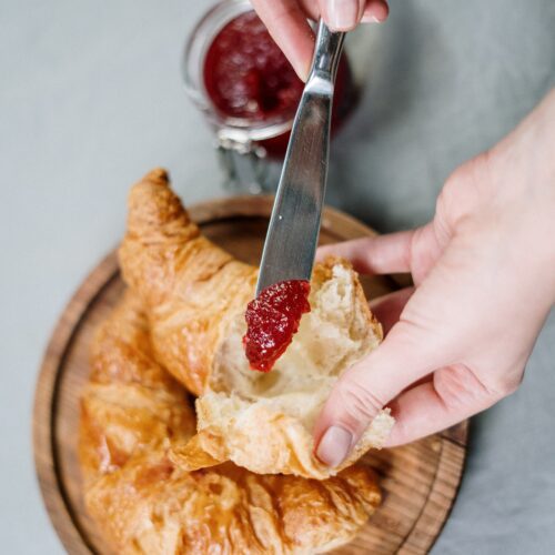 Hands holding a torn open croissant with a knife with some redcurrant jelly on it, ready to be smeared onto the crioissant. Jar/bolw of redcarrant jelly in the background