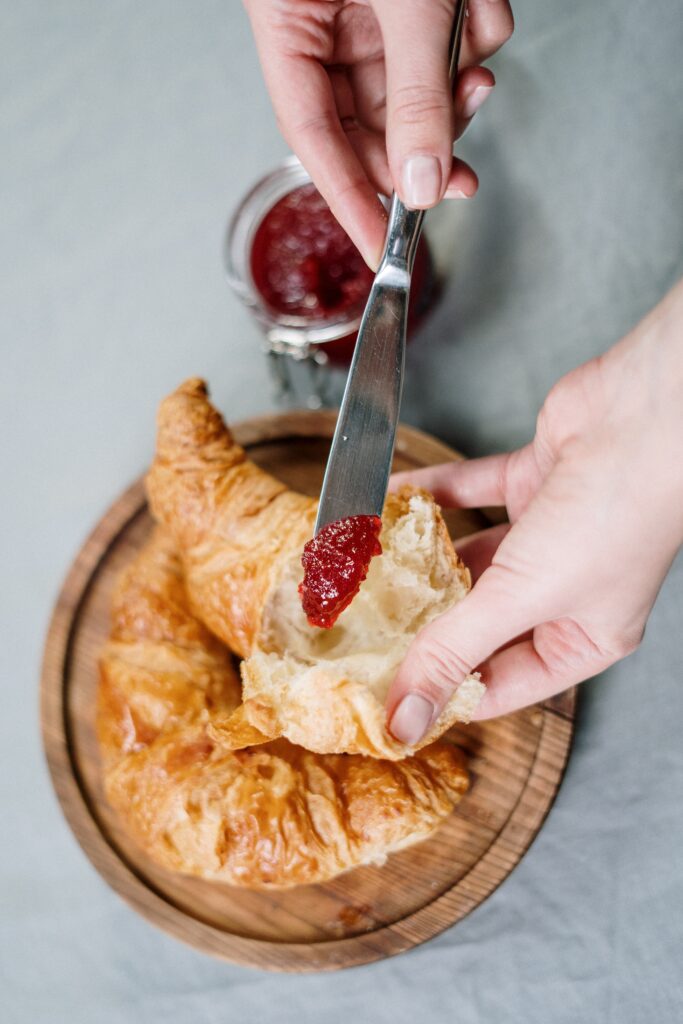 Hands holding a torn open croissant with a knife with some redcurrant jelly on it, ready to be smeared onto the crioissant. Jar/bolw of redcarrant jelly in the background