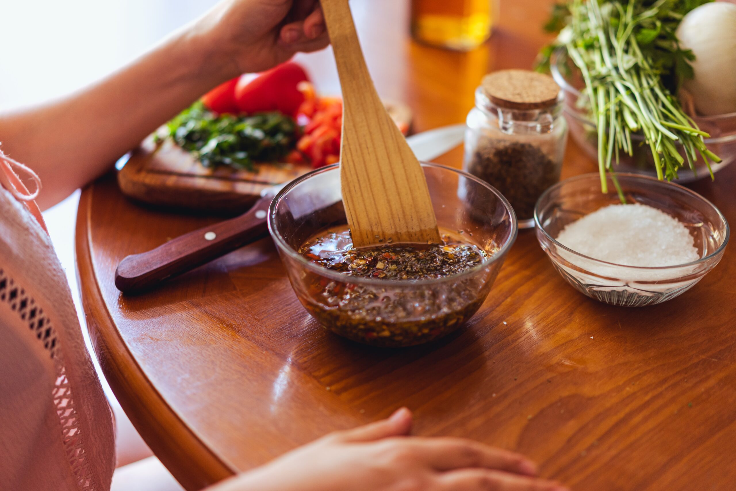 Womans hands holding a wooden spoon stirring a spicy mixture in a glass bowl. Chopped vegetables and a jar of sices on the work-surface in the background