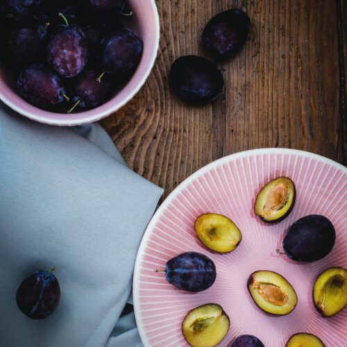 A bowl of plums on a table. Bowl standing next to plums cut in half with halves facing upwards on plate and with stones in place