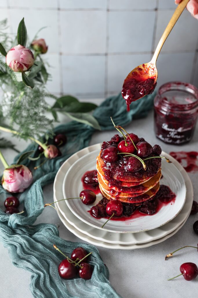 Cherry compote being drizzled over a stack of pancakes on a plat. Jar of compote, peonies and green leaves on table in the background.