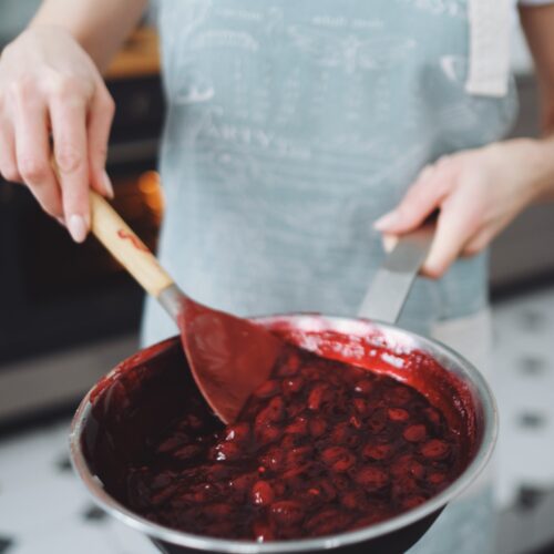 Person wearing a light grey apron, stirring a pan full of cherries in syrup with a wooden spoon.