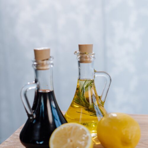 2 cork stopperred pouring bottles, one with oil and one with blackberry vinegar. They are standing on a wooden board, with one whole and one cut lemon in the forground.