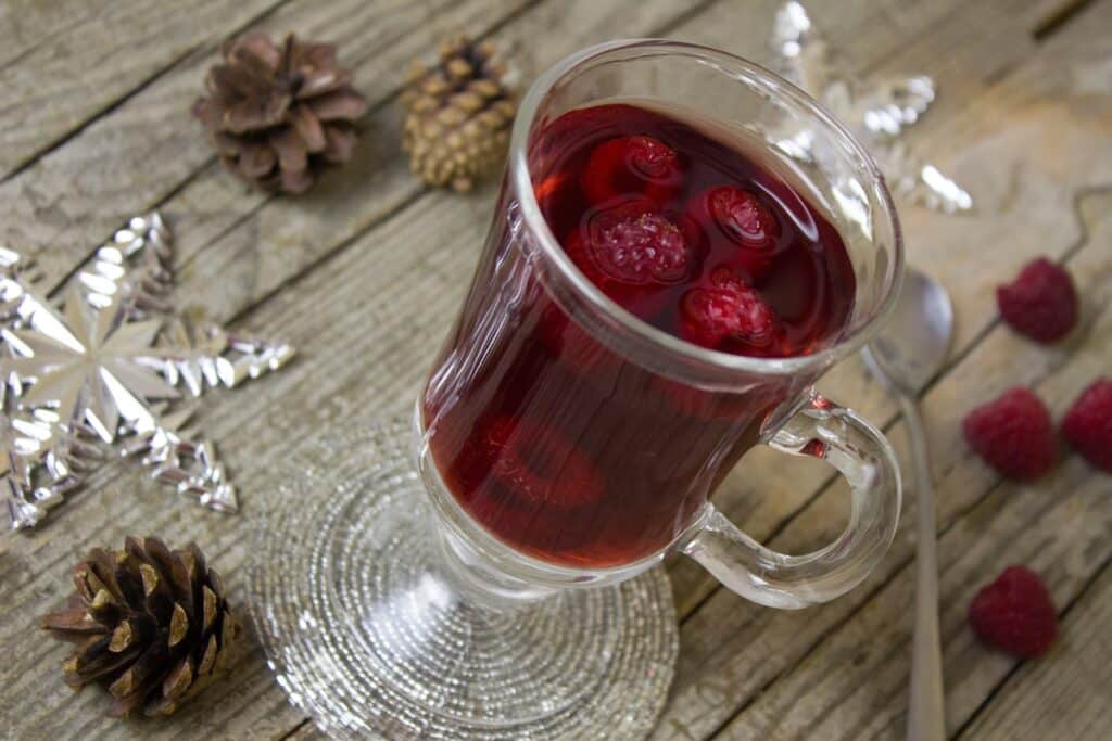 A stemmed glass mug of raspberry cordial with whole raspebrries floating in it. Standing on wooden table and some acorns in the background.