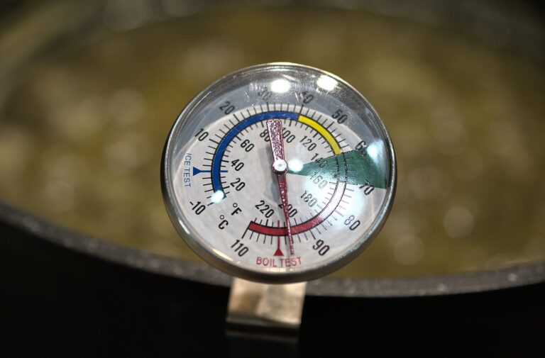 Dial of a sugar thermometer attached to the edge of a pot. Boiling jam in the blurred background.