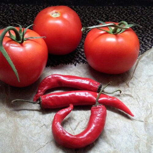 3 red tomatoes on a mostly white, and black background. With 3 red chillies in the foreground in front of the tomatoes.