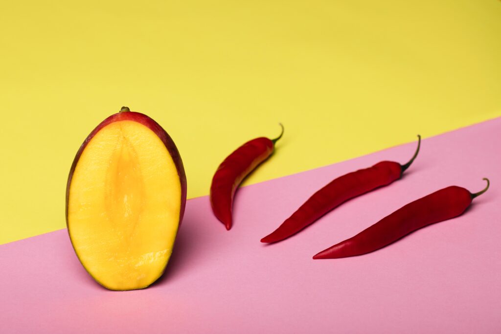 Yellow wall and pink surface. Whole mango with a slice taken showing the yellow flesh, and next to it, three red chillies lined up in a diagonal row.