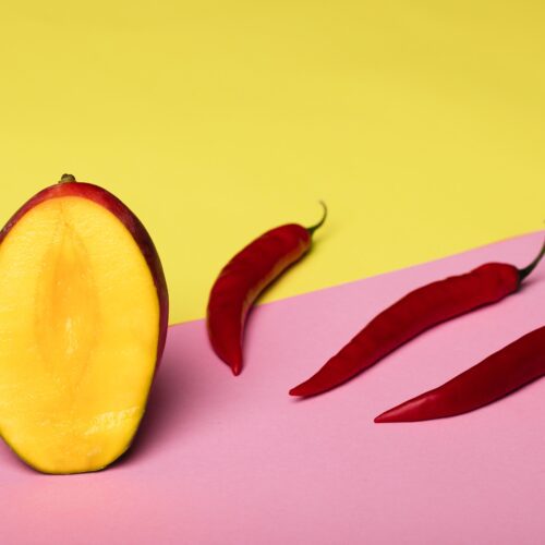 Yellow wall and pink surface. Whole mango with a slice taken showing the yellow flesh, and next to it, three red chillies lined up in a diagonal row.