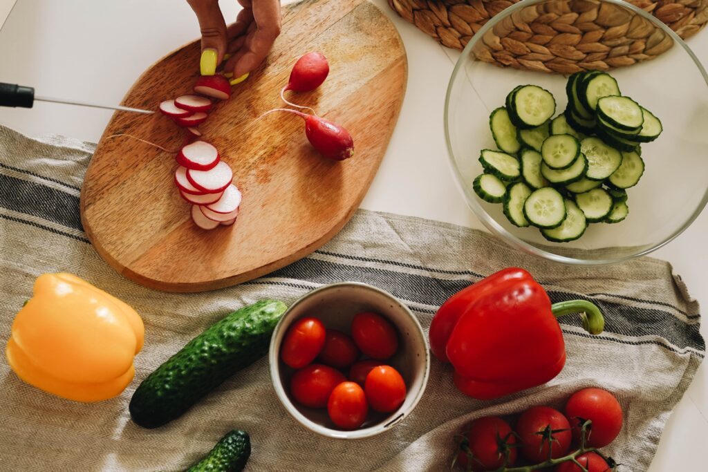 A person preparing vegetable, chopping radishes on wooden board with other vegetables surrounding it.