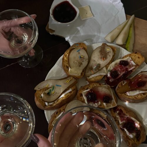 Platter of savoury bites with slices of pickled pears on bruschetta. Hands holding glasses of sparkling above the platter.