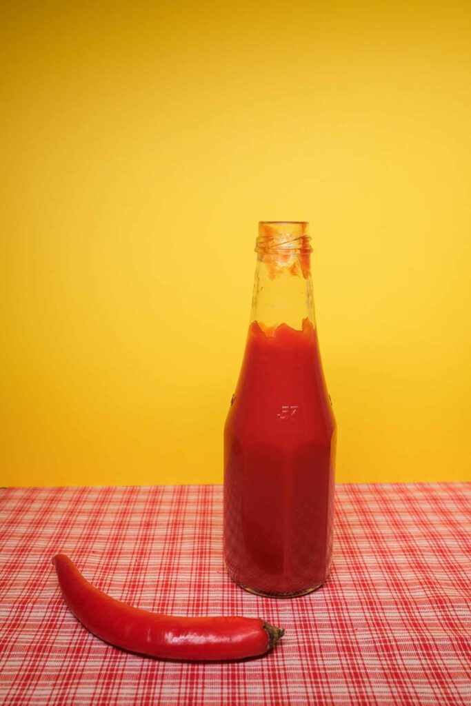 Lidless bottle of chilli ketchup standing on a red and white checked tablecloth with a large red chilli in front of the bottle.