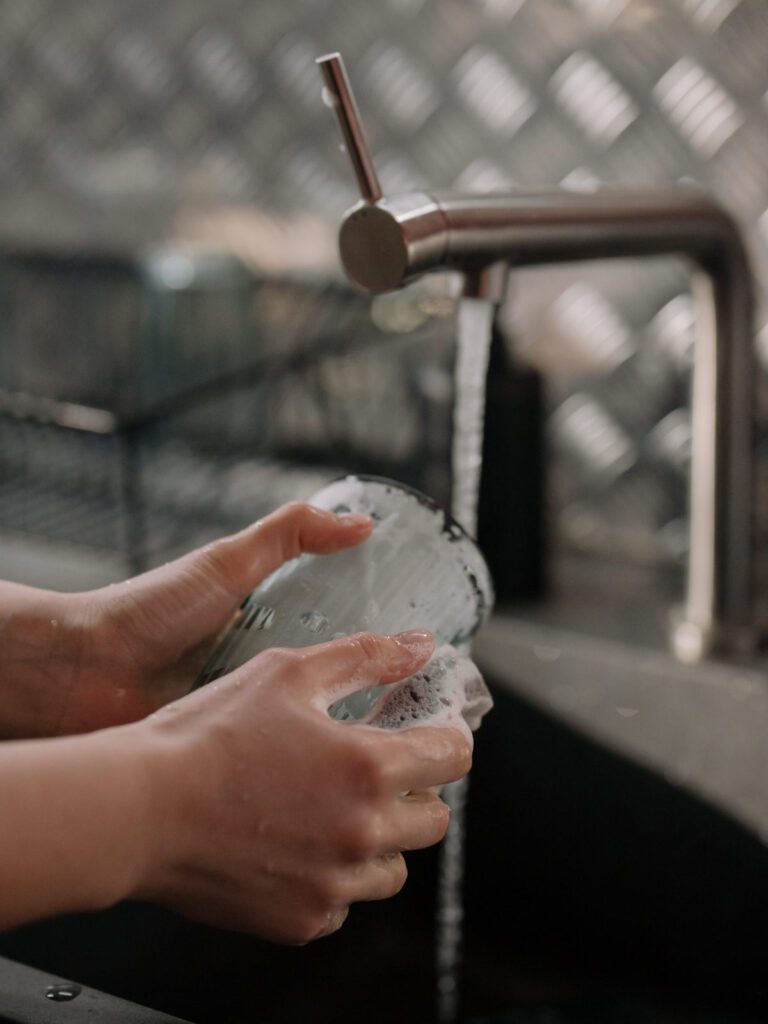 Person washing glass at sink with running water and foamy soap.