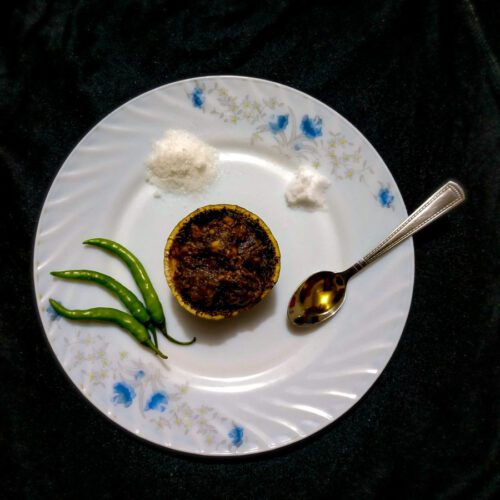 A small bowl of rhubarb and date chutney in the middle of a white and blue-floral edged plate. A single spoon, three green chillies and two small heaps of salt on the plate surrounding the bowl of chutney.