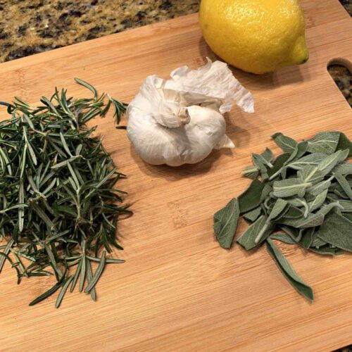 Chopped rosemary, a bulb of garlic a lemon and more fresh herms on a chopping board.