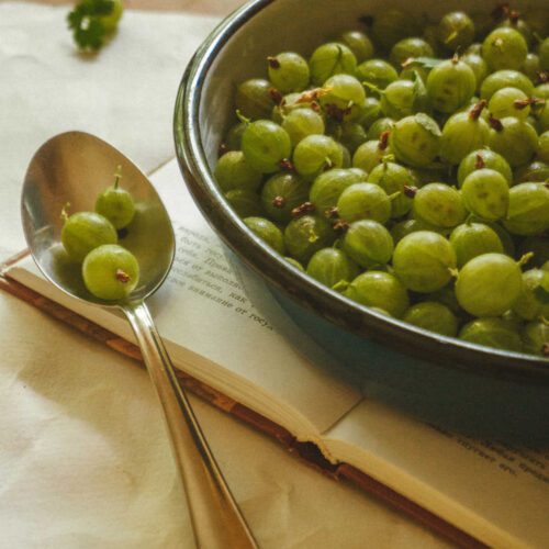 Gooseberries in a shallow bowl resting on a recipe book. Spoon with a few gooseberries in it to the left of the bowl.