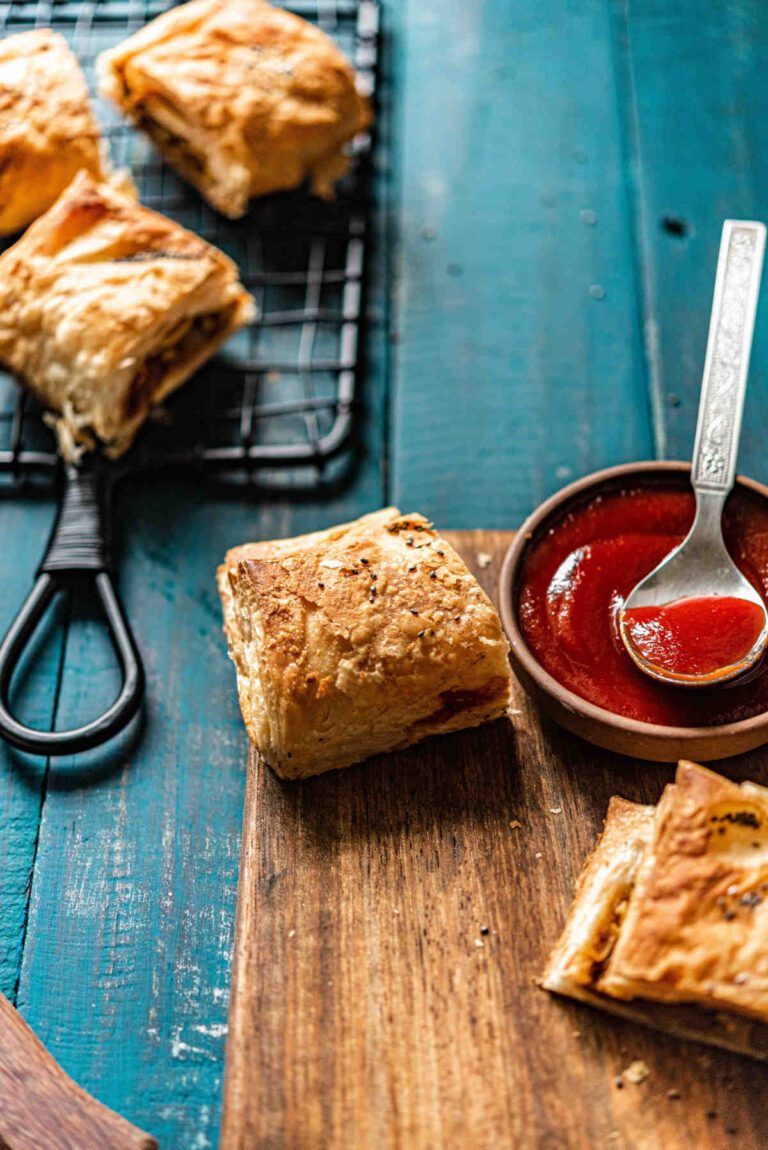 A pot of bloody mary ketchup with a spoon on a wooden board. Sausage rolls on tray and on board.