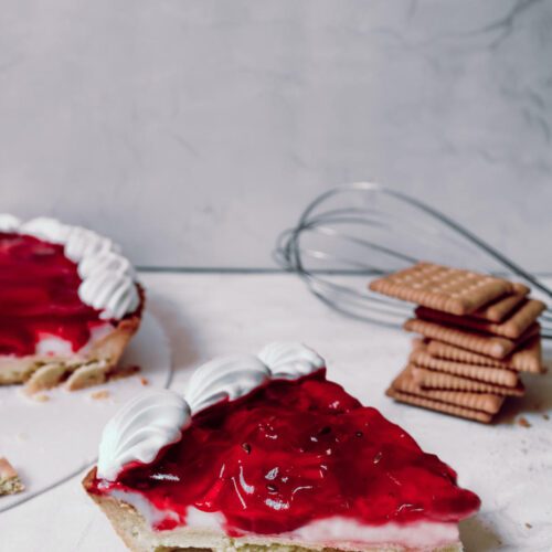 A slice of a desert tart topped with strawberry gooseberry jam. The rest of the tart in the background alongside a pile of plain busicuits.