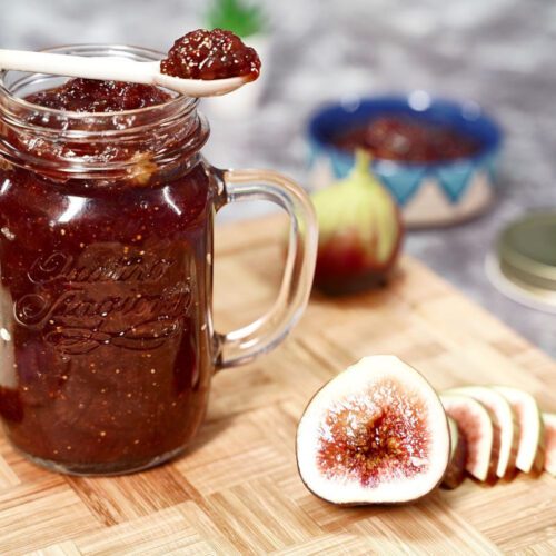 An open jar of fig and ginger jam on a wooden chopping board with cut sliced ripe fig in the foreground.