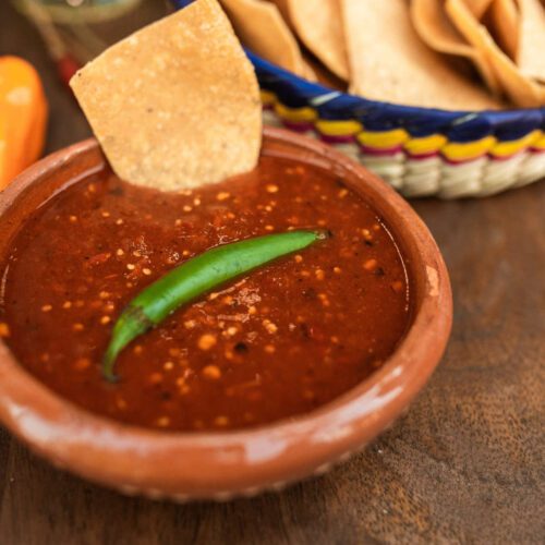 A terracotta bowl of garlic hot sauce with a green chilli garnish, standing next to a bowl of tortilla chips - one chip struck into sauce.