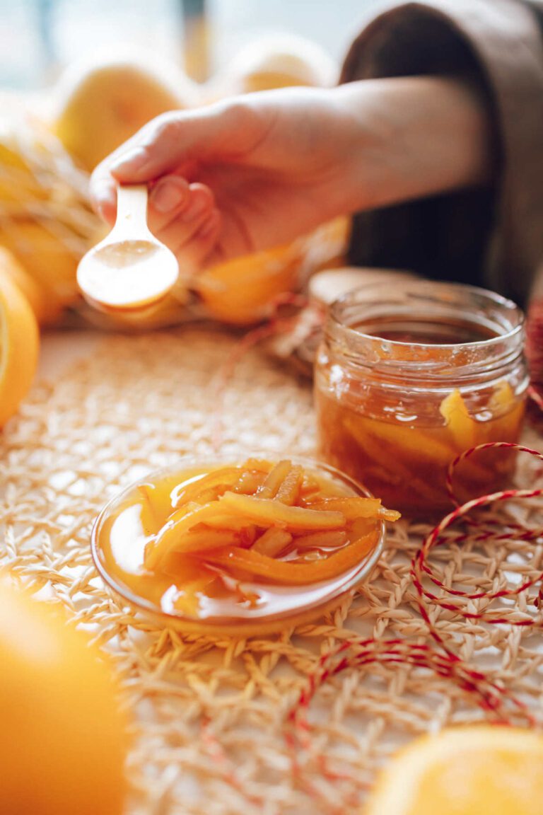 A hand holing a wooden spoon having spooned out some runny orange marmalade onto a glass plate. A jar of the marmalade stands alongside.