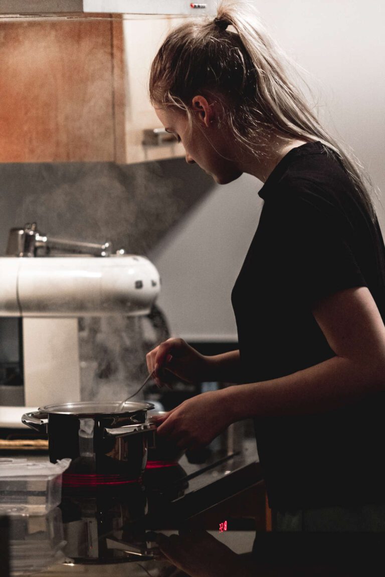 A woman cooking and stirring a pot on the hob. Steam rising from it. Kitchen cupboard and stand mixer in the background.