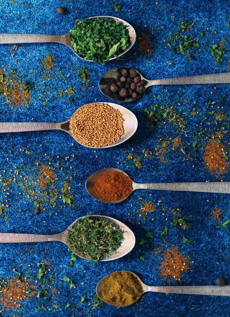 Metal spoons filled with different spices and herbs on a blue background.