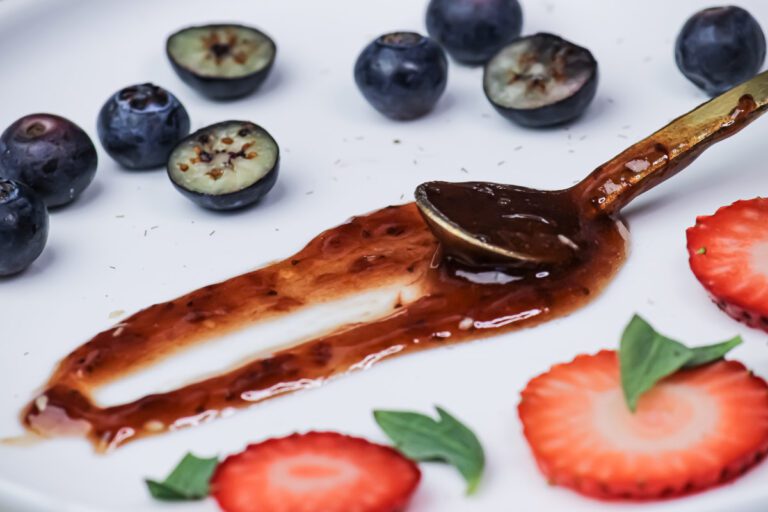 A small spoon dragged through a pool of dark jam on a white plate, and leaving a trough. Cut strawberries and blueberries in the foreground and background.