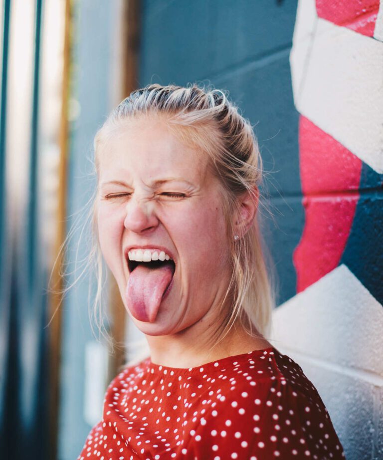 A blonde haired woman with face scrunched up, eyes closed and tongue out as it having tasted something unpleasant.