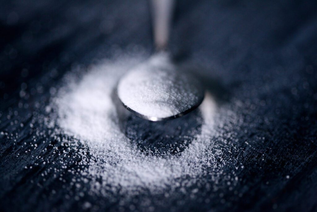 A close up image of fine caster sugar in a spoon with some spilled around the spoon onto a dark surface.