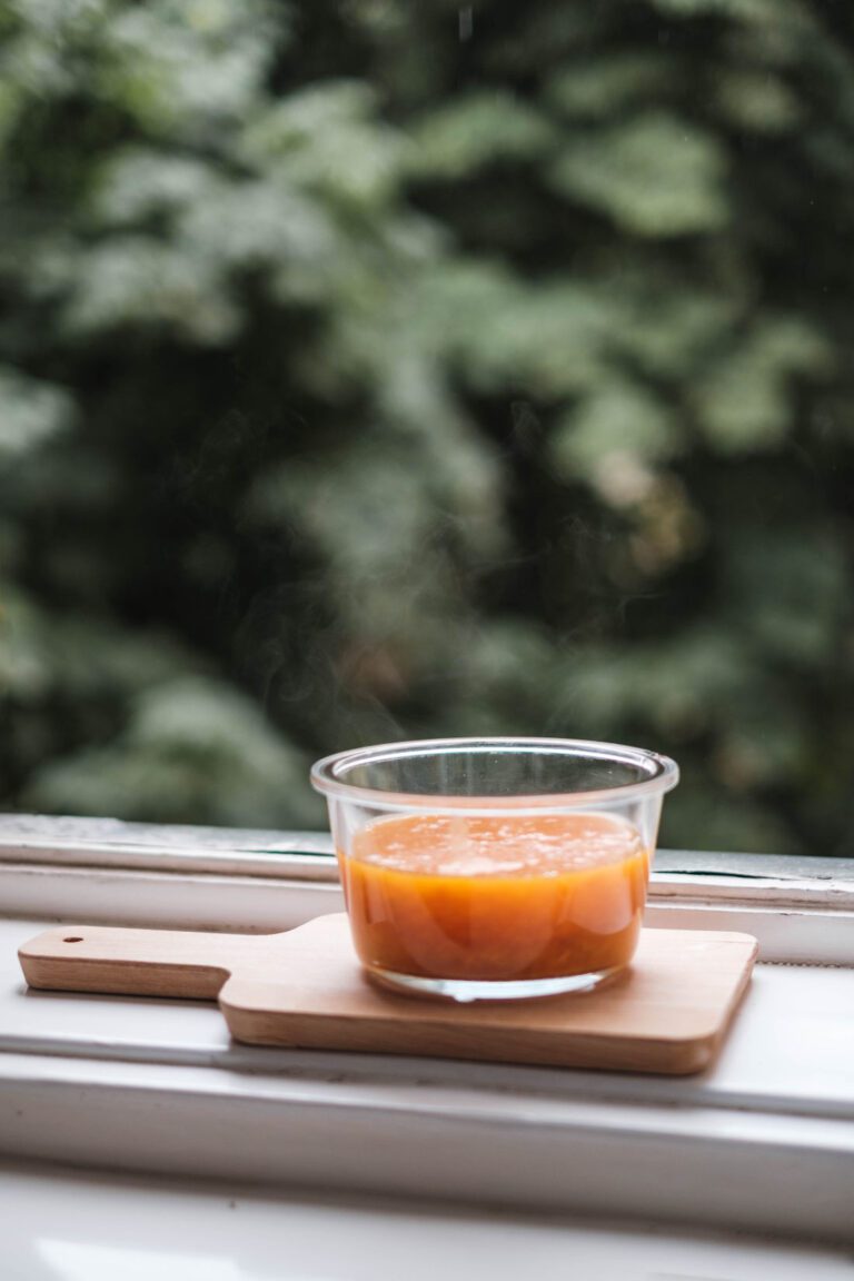 A glass pot of mango chutney on a small wooden paddle, on a white windowsill.