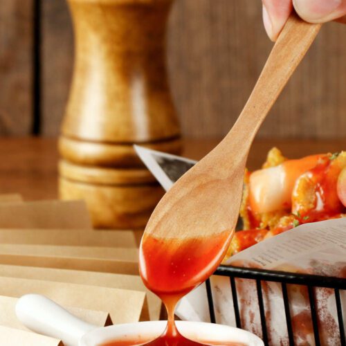 Mango chutney in a white pit - a wooden spoon has been dipped into the sauce and is raised with sauce dripping from it. A basket of fried food in the background.