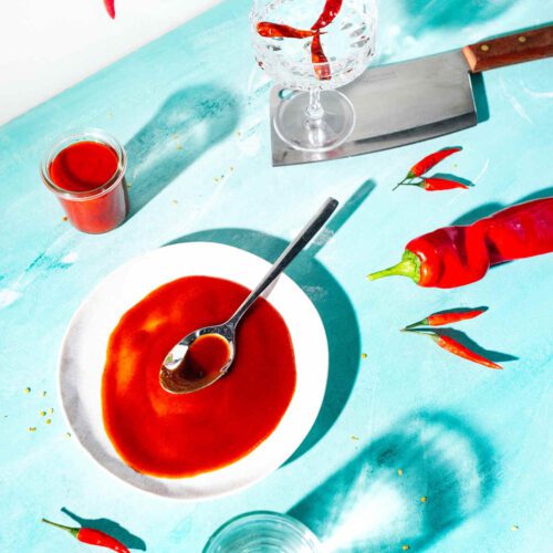 Tomato ketchup on a plate with a spoon, in a small pot alongside. Chillies around it, a knife and glass of clear liquid.